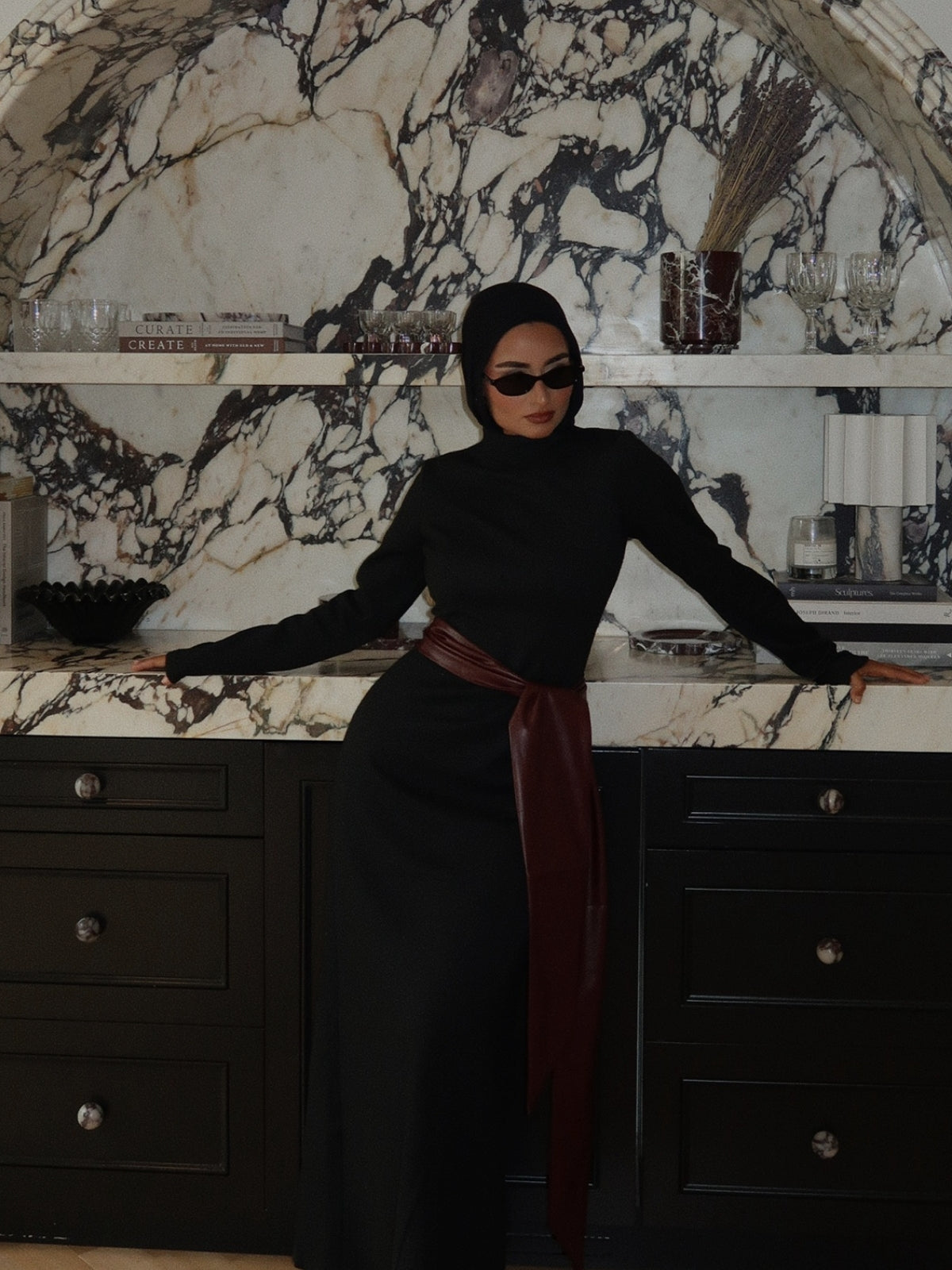 Woman in a black hijab and black maxi dress with a burgundy wrap belt, leaning on a marble counter