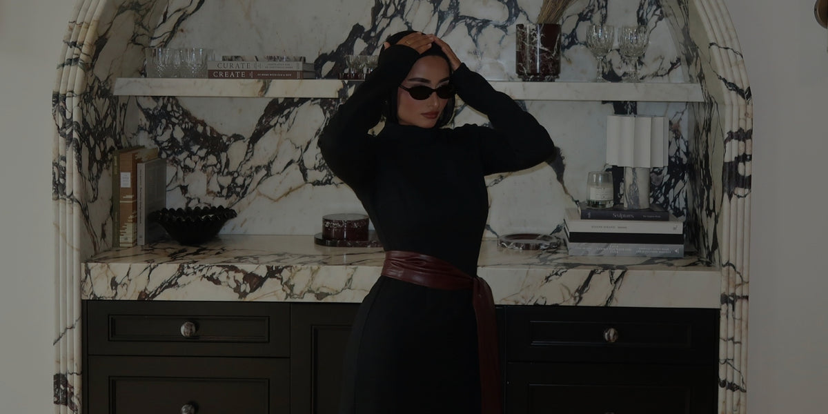 Woman in a black hijab, sunglasses and black maxi dress posing in front of a marble arched shelf for the VEILED Annual Members Sale